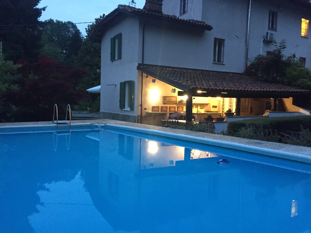 An inviting outdoor area is highlighted by a clear, blue swimming pool surrounded by greenery. The entrance to an open-plan living space is visible, with soft lighting illuminating the interior. The building's classic architecture features large windows and a tiled roof.