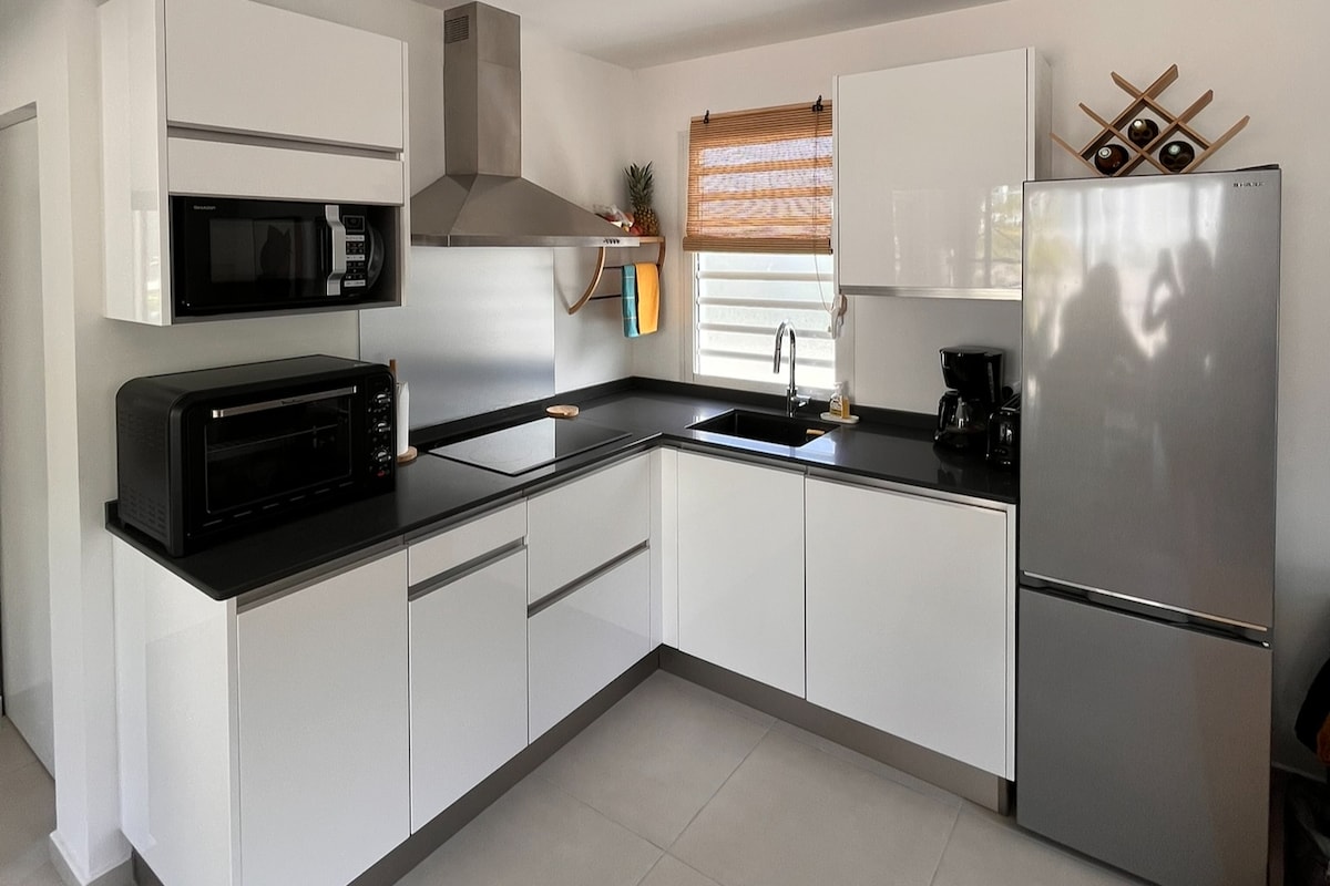 A modern kitchen features sleek white cabinetry and contrasting dark countertops. Stainless steel appliances, including a microwave, refrigerator, and coffee maker, are neatly arranged. Natural light enters through a window adorned with bamboo blinds, highlighting the organized workspace.
