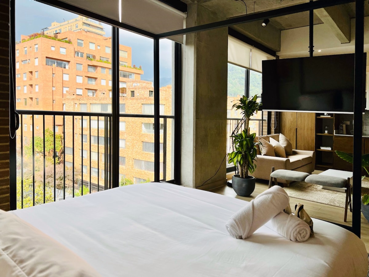 A bright sleeping area features a spacious bed with crisp white linens and neatly arranged towels. Large windows provide views of the surrounding cityscape and greenery. A comfortable seating area is visible, enhancing the sense of spaciousness and natural light within the loft.