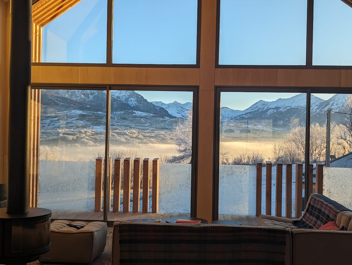 Large windows provide a panoramic view of surrounding mountains and valleys, which are partially shrouded in mist. A cozy seating area is visible in the foreground, with natural light filling the space and highlighting the wooden accents throughout.