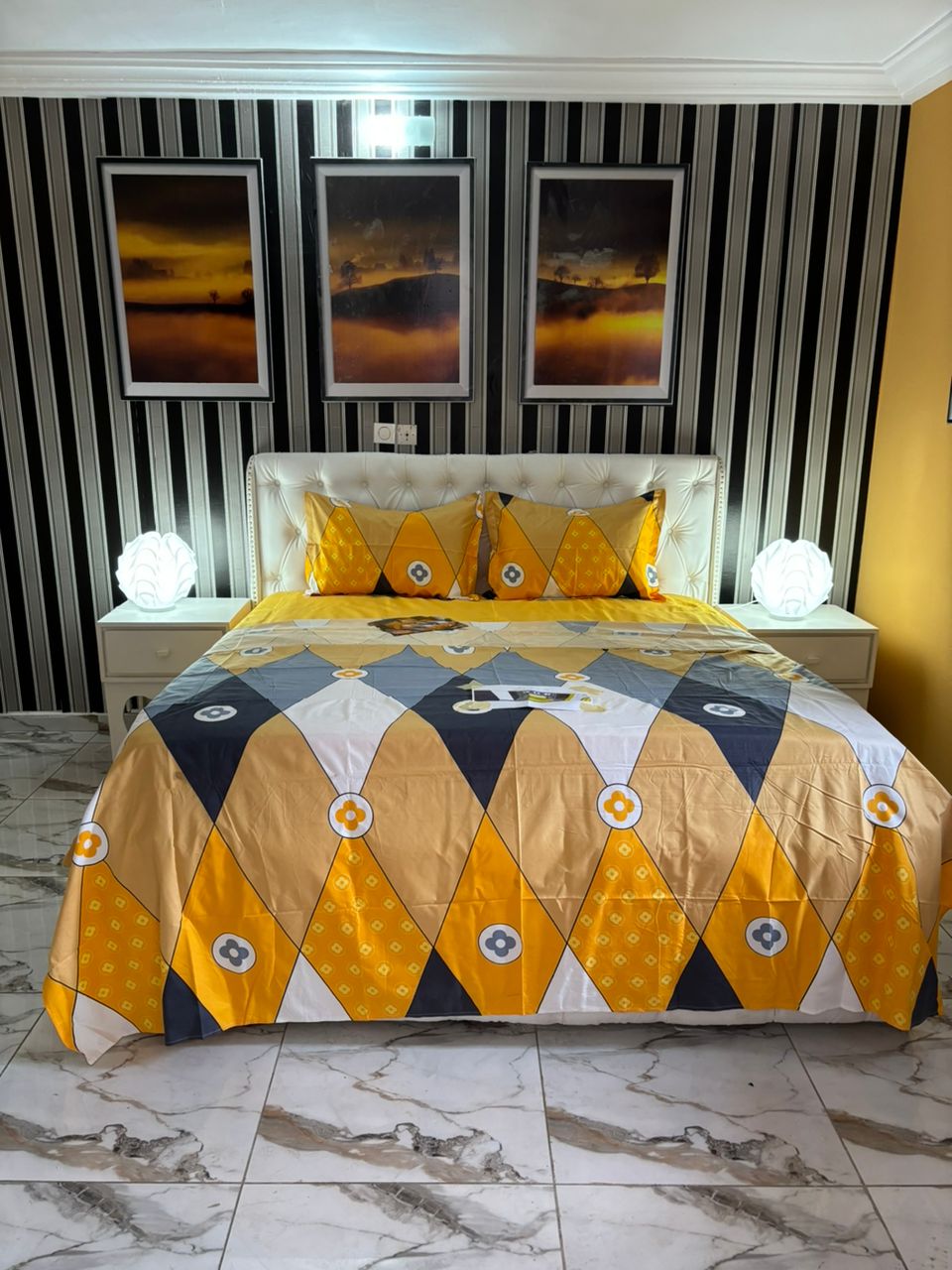A sleeping area is presented with a light-colored upholstered bed adorned with a bold, patterned bedding set featuring geometric shapes in yellow, gray, and white. Two complementary pillows sit atop the bed. Soft lighting is provided by two bedside lamps, and artwork hangs on the striped wall.