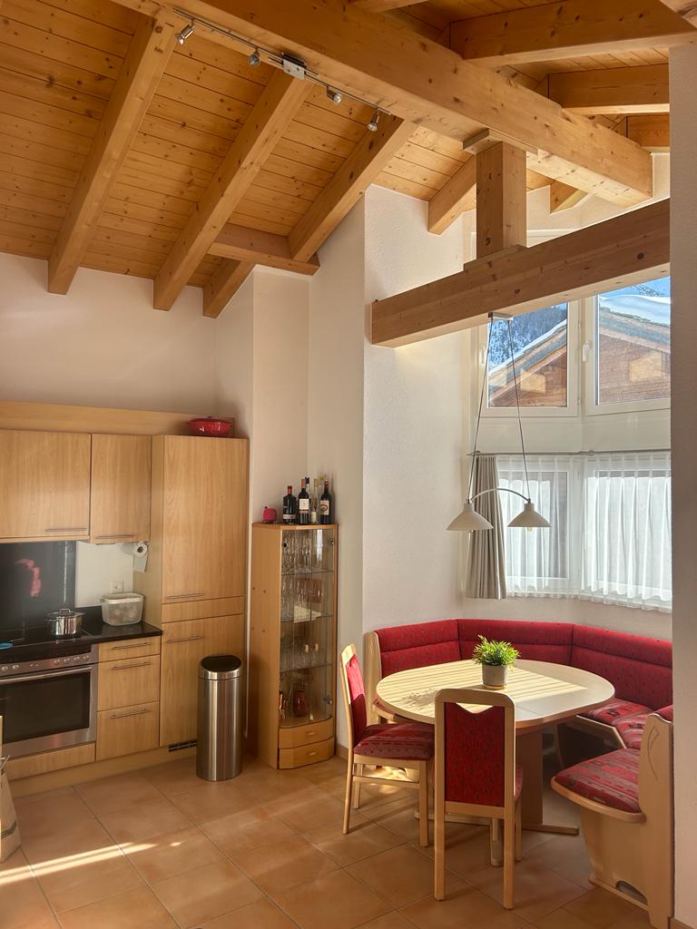A bright kitchen area features wooden beams overhead, with natural light filtering through large windows. A round dining table is surrounded by red cushions, while wooden cabinetry lines one wall. Nearby, a glass-fronted cabinet displays various items, enhancing the inviting atmosphere.