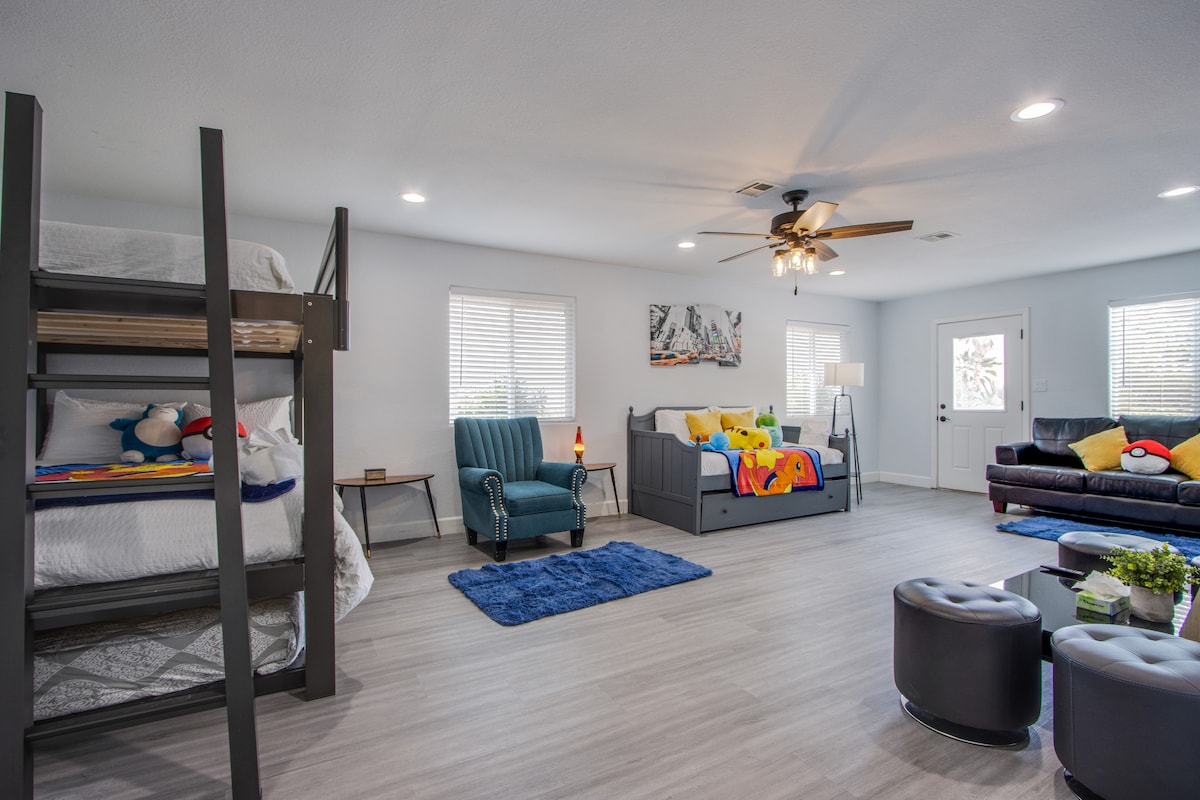 A spacious common area is designed for relaxation and comfort. A bunk bed with colorful bedding stands alongside a daybed adorned with bright throw pillows. A cozy armchair and a sofa with vibrant cushions create inviting seating options, illuminated by natural light through multiple windows.
