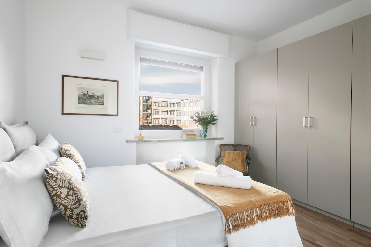 A serene bedroom features a spacious bed with decorative pillows and a soft throw at the foot. A large wardrobe is positioned against the wall, and natural light fills the room through a window, offering a view of the cityscape outside.