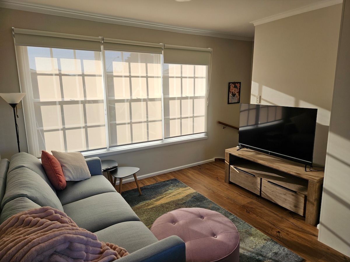 A bright living space features a light-toned sofa adorned with cushions, complemented by a round ottoman in soft pink. A wooden TV unit holds a flat-screen television, while large windows allow natural light to fill the room, creating an inviting atmosphere.