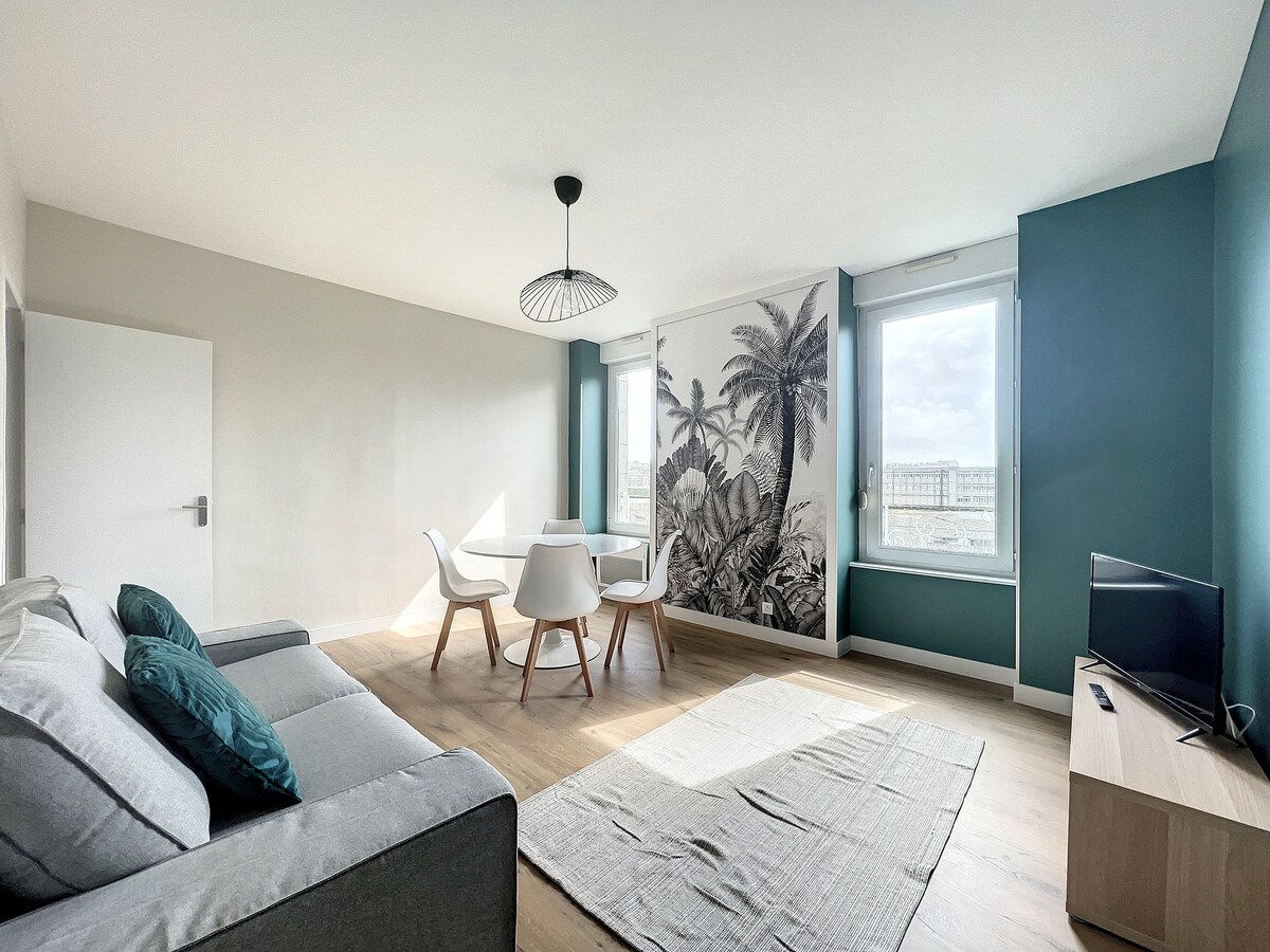 A modern living area features a comfortable gray sofa and a small wooden television stand. A dining table with white chairs is positioned near windows that allow natural light to illuminate the space. Green walls create a calming atmosphere, complemented by a large mural of tropical foliage.