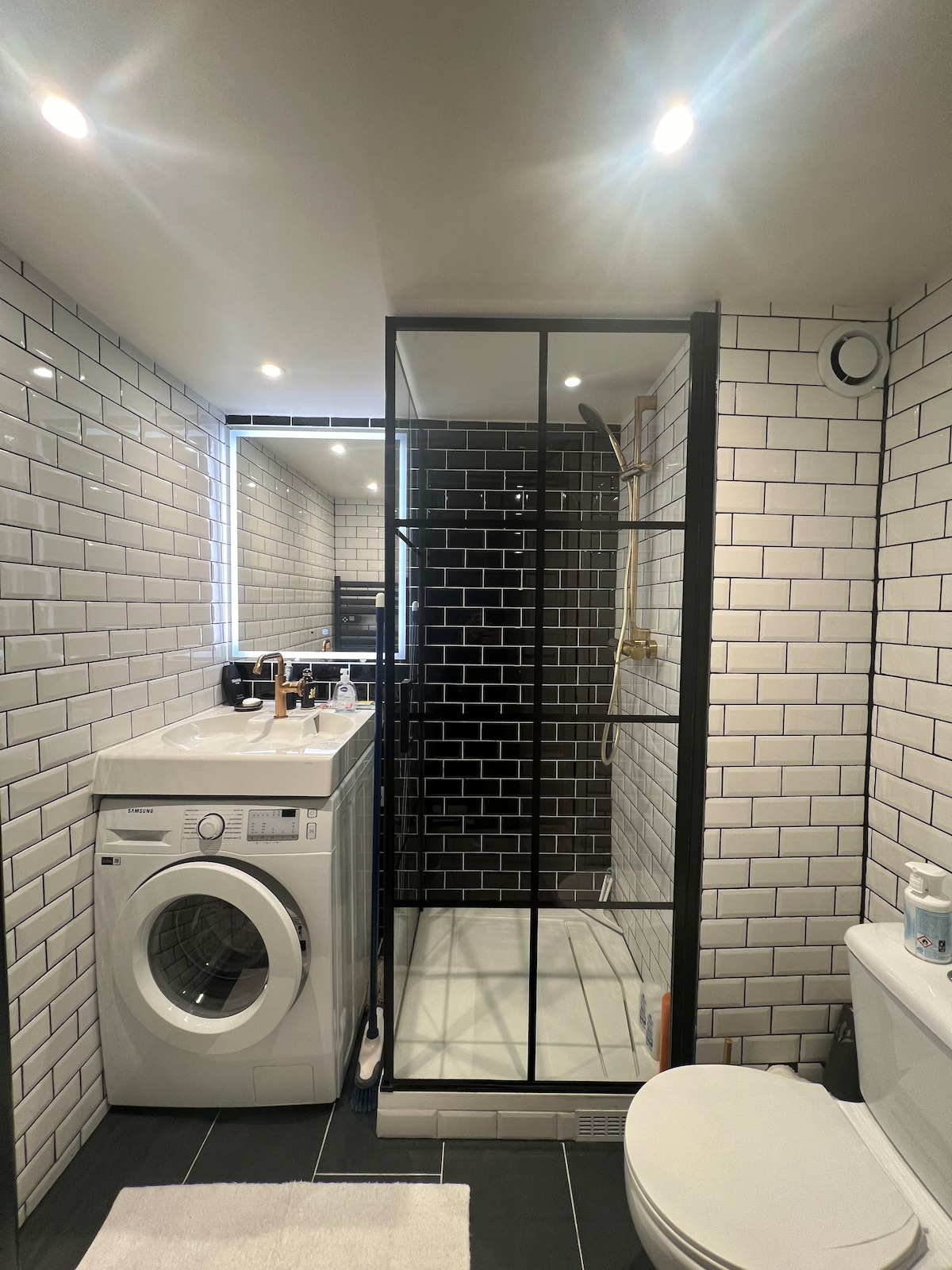 A modern bathroom space features sleek black and white subway tiles throughout. A glass shower enclosure is situated next to a washing machine. Ample lighting is provided by recessed ceiling lights, while a white bath mat is placed on the floor near the toilet.