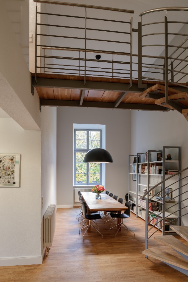 A spacious dining area is illuminated by natural light from a large window. A long wooden table is surrounded by black chairs, while a dark pendant light hangs overhead. A staircase with a metal railing leads to the upper level, and a bookshelf displays various items.