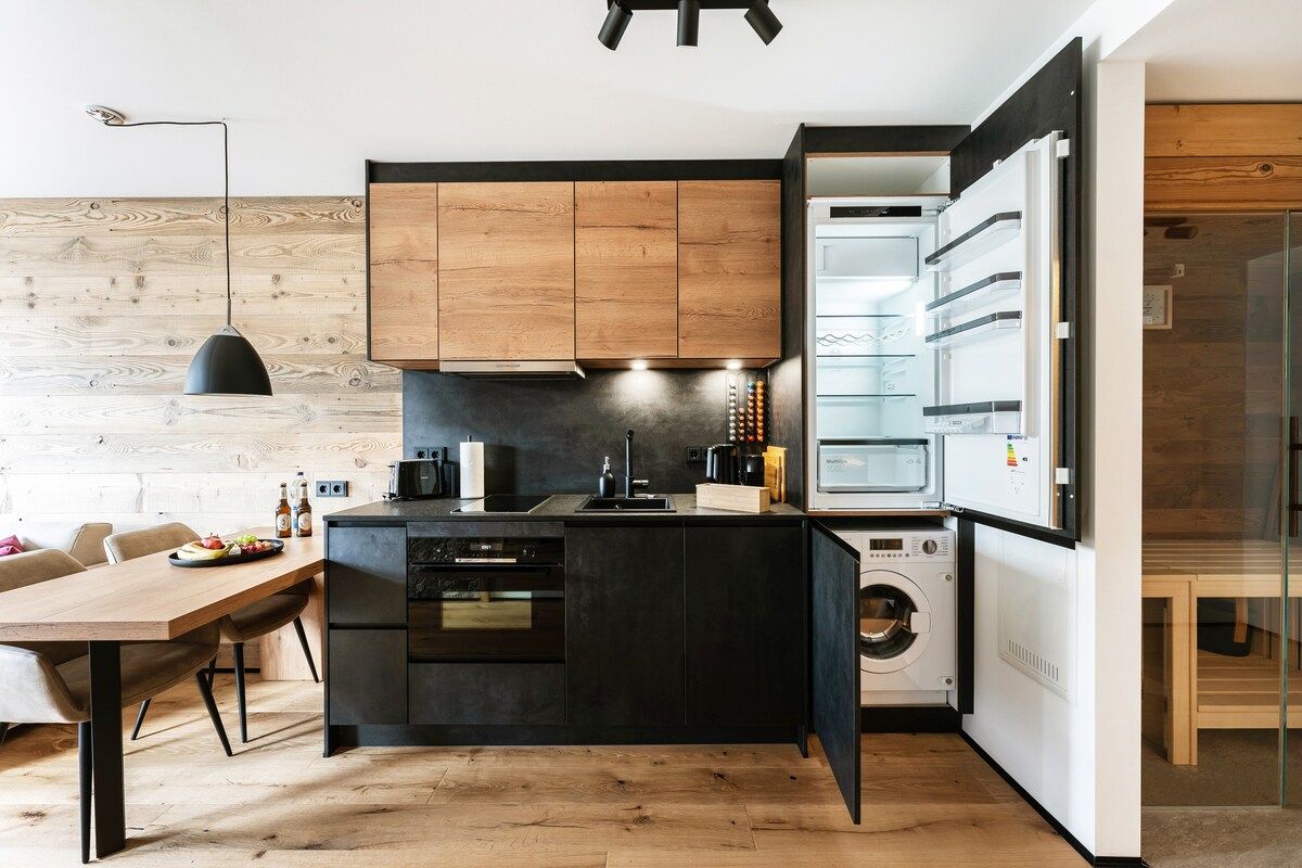 A modern kitchen is presented with sleek black cabinetry and warm wooden accents. Equipped appliances include a built-in oven, stovetop, and a refrigerator with a glass door. A wooden dining table is visible nearby, accompanied by soft seating and decorative elements.
