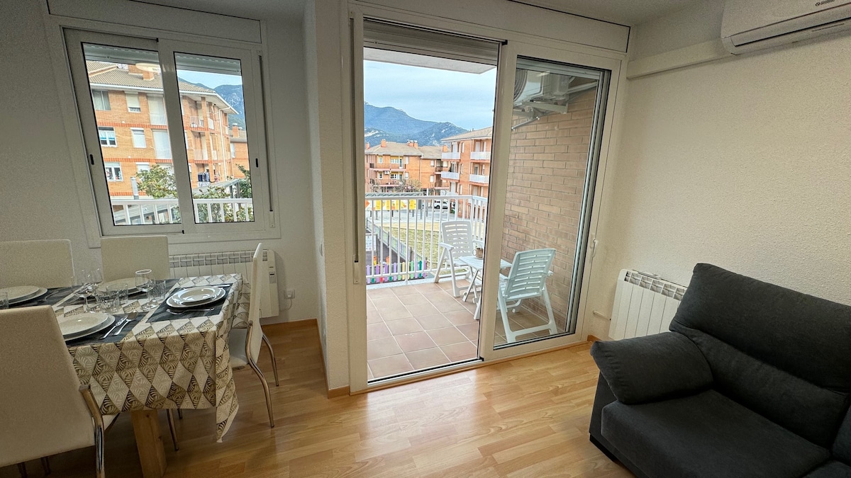 A bright living area features a comfortable dark gray sofa and a dining table set for four. Large glass doors open to a balcony with two outdoor chairs, offering views of surrounding buildings and distant mountains.