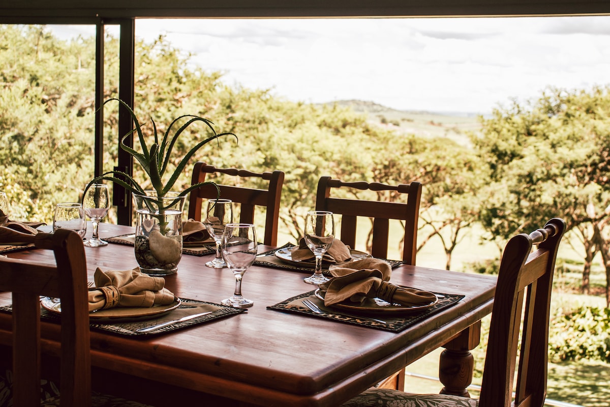 A wooden dining table is set for six, featuring simple yet elegant tableware. Softly folded napkins and glasses are arranged on placemats. Behind the table, large windows reveal views of lush greenery and the distant landscape.