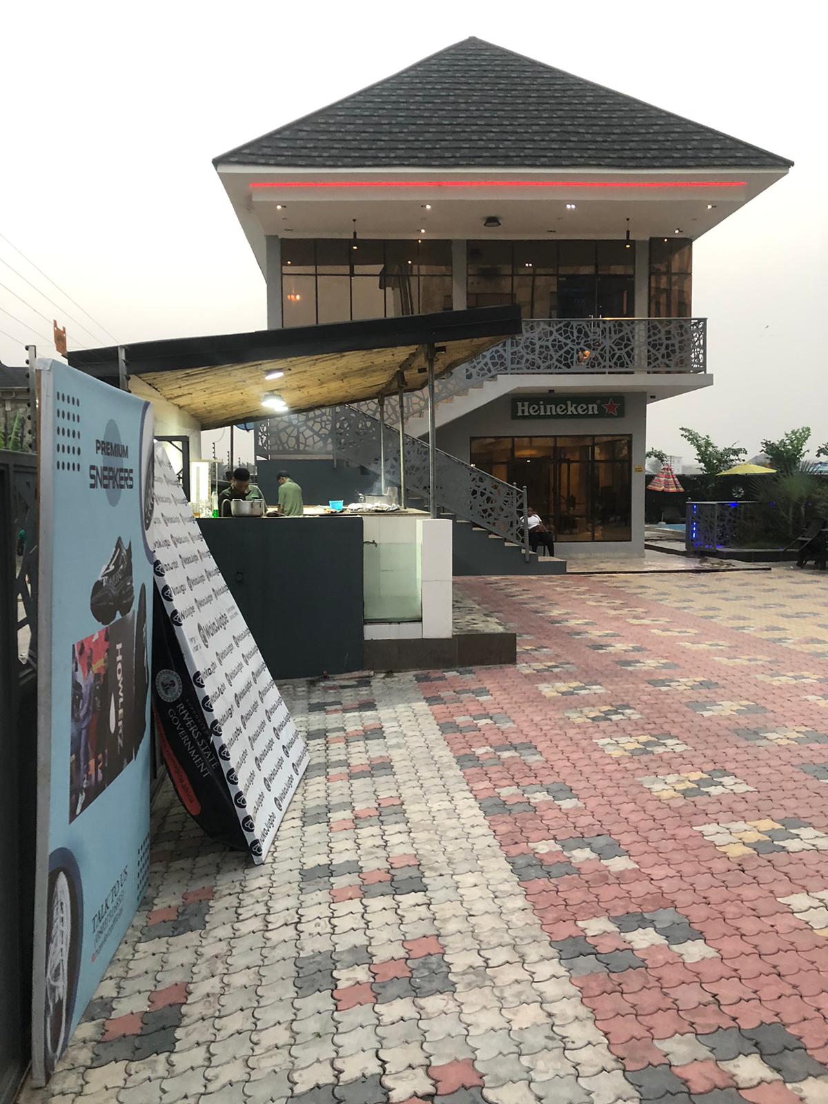 A modern two-story building with a sloped roof is shown. An outdoor seating area is visible, with colorful pavers leading towards the entrance, where a sign indicates a Heineken establishment. Green plants add natural elements to the inviting outdoor space.