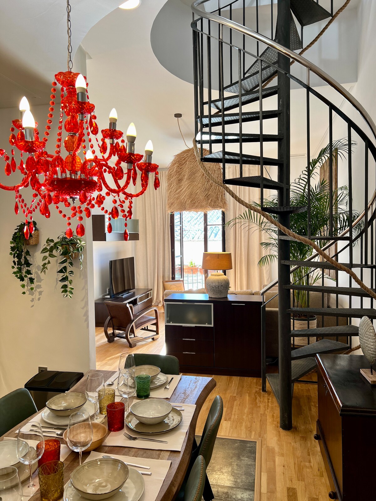 A dining area features a wooden table set for six, complemented by colorful glassware. A vibrant red chandelier hangs above, illuminating the space. A spiral staircase leads to the upper level. Natural light filters through a window, creating a cozy atmosphere in the room.