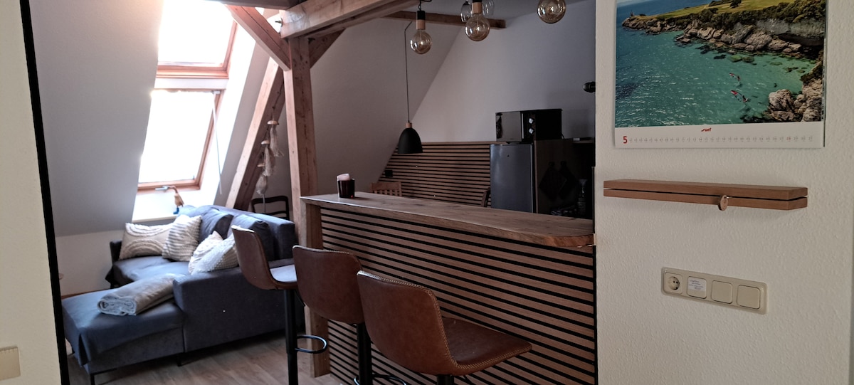 A contemporary living area features a cozy sectional sofa alongside a wooden bar with high-backed stools. Light streams through the skylights, illuminating the warm wood tones. An elegant black pendant light hangs over the bar, while a nature-themed calendar adorns the wall.
