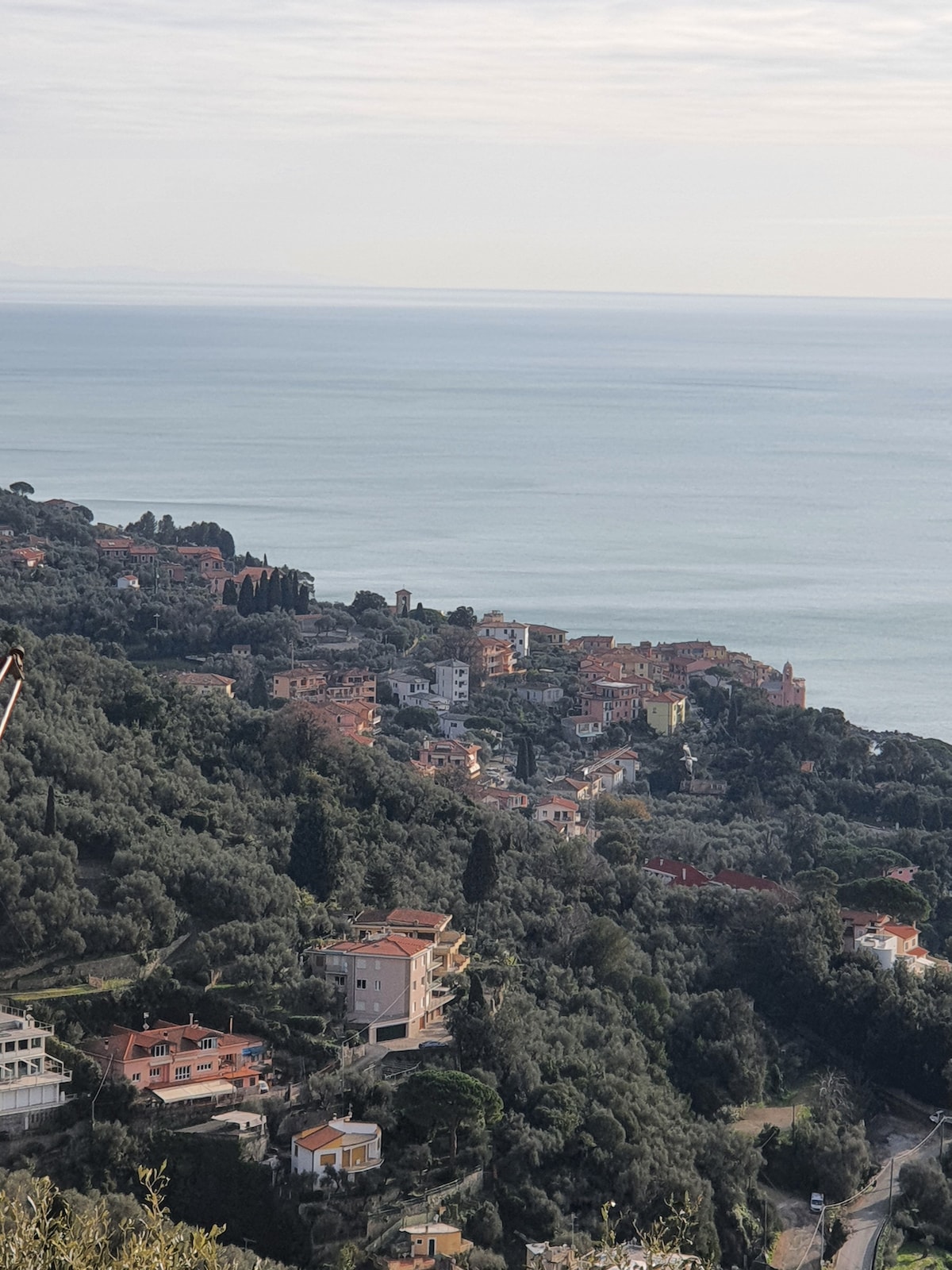The image captures a panoramic view of a coastal village nestled among rolling hills. Colorful residential buildings are interspersed with lush greenery, while the serene sea stretches into the horizon under a light sky.