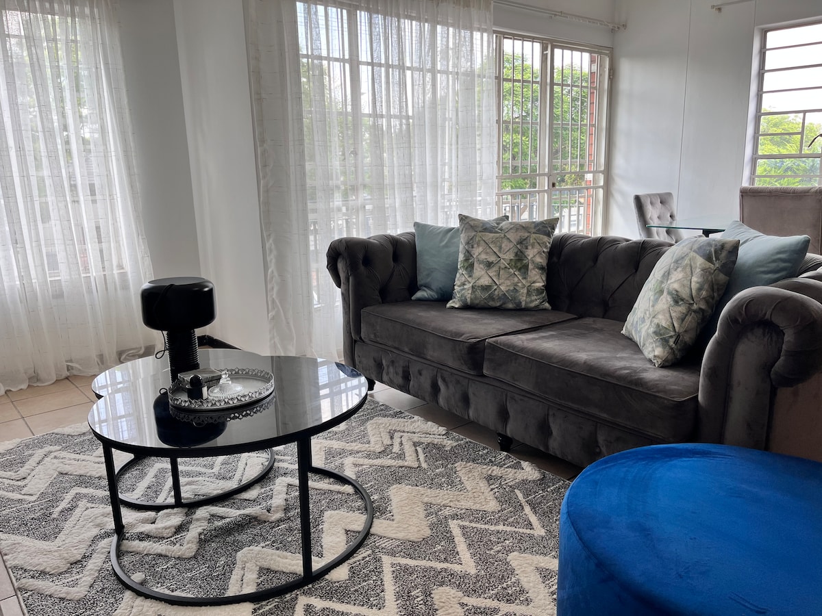 A modern living area is featured with a plush gray sofa adorned with patterned cushions. A round black coffee table sits on a patterned rug, while natural light enters through large windows draped with sheer white curtains.