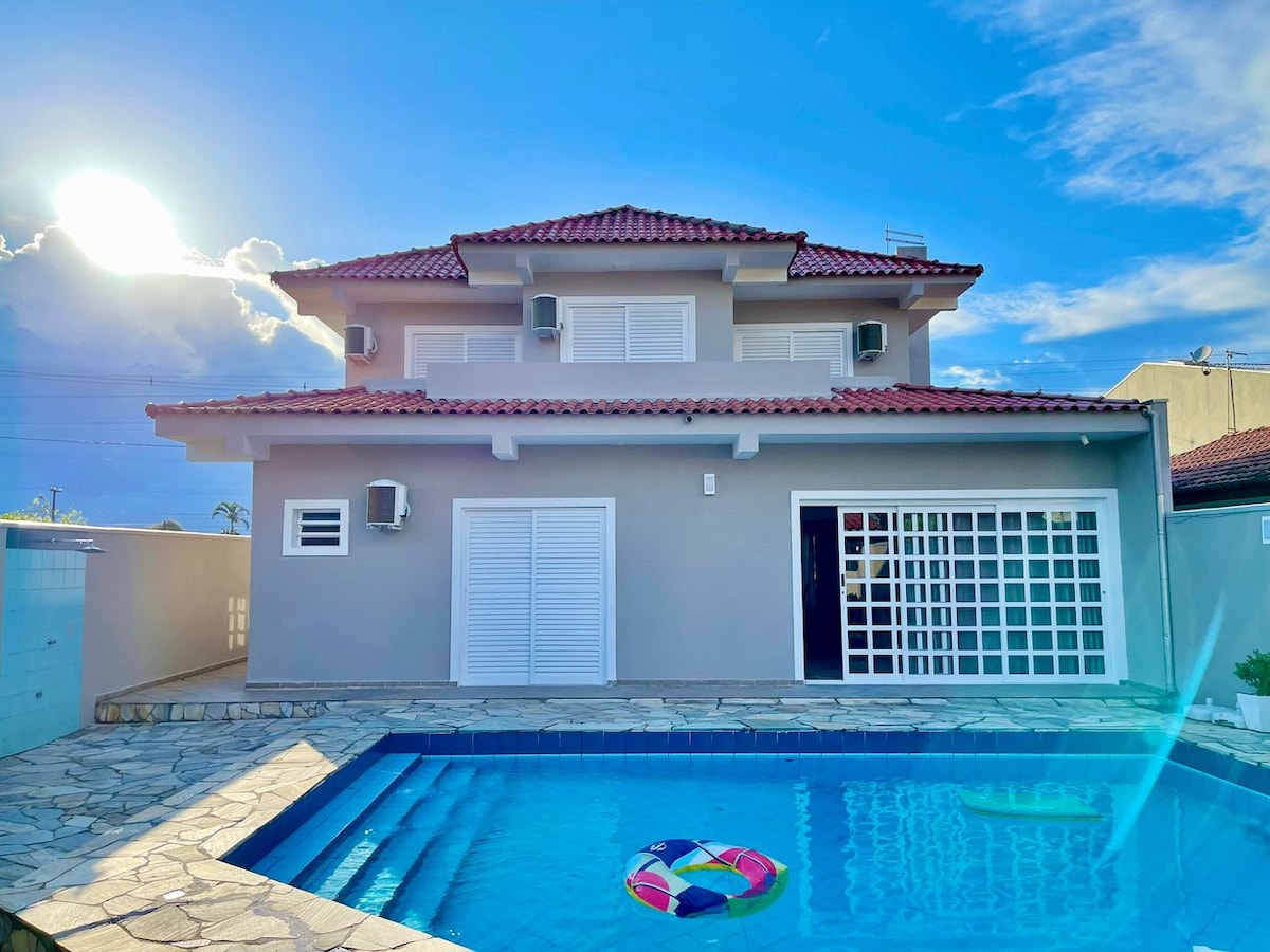 A two-story house is depicted, featuring a red-tiled roof and multiple air conditioning units. The pool area is visible, along with a stone deck and colorful pool floats. Sunlight is seen illuminating the home, creating a bright and welcoming exterior.