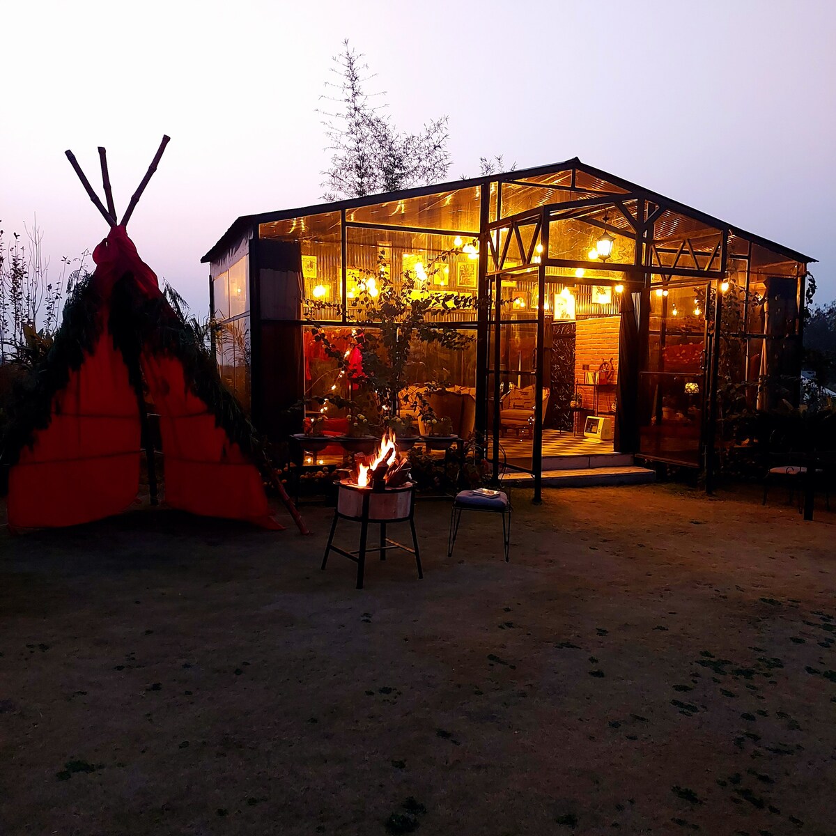 A cozy outdoor cabin is surrounded by a green lawn, illuminated by warm lights. At the forefront, a small fire burns in a metal container near a chair. A decorative, red teepee structure is positioned nearby, enhancing the outdoor ambiance.