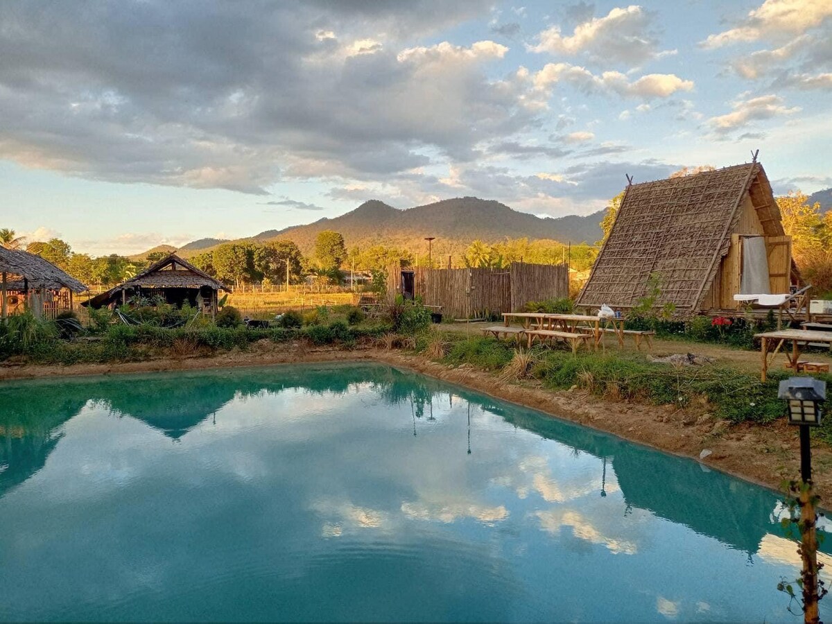 A tranquil landscape is depicted, featuring a mirror-like pond reflecting the sky and nearby mountains. Two traditional huts are positioned near the water's edge, surrounded by lush greenery and wooden seating areas that invite relaxation.