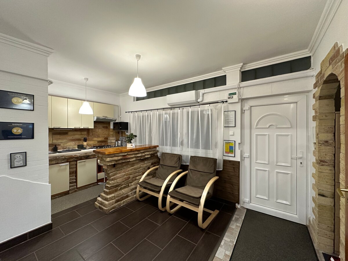 An entrance area is shown, featuring two wooden chairs aligned against a wall. A counter made of stone is visible, while the kitchen area is seen in the background. Soft lighting is provided by ceiling lamps, and a white door leads to another space.