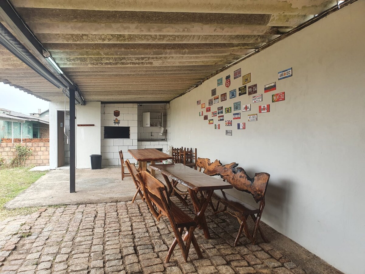 An outdoor dining area is featured under a covered patio. Several wooden tables, including one with a natural edge, provide ample seating. A collection of decorative signs adorns the wall, while a simple outdoor kitchen can be seen in the background.