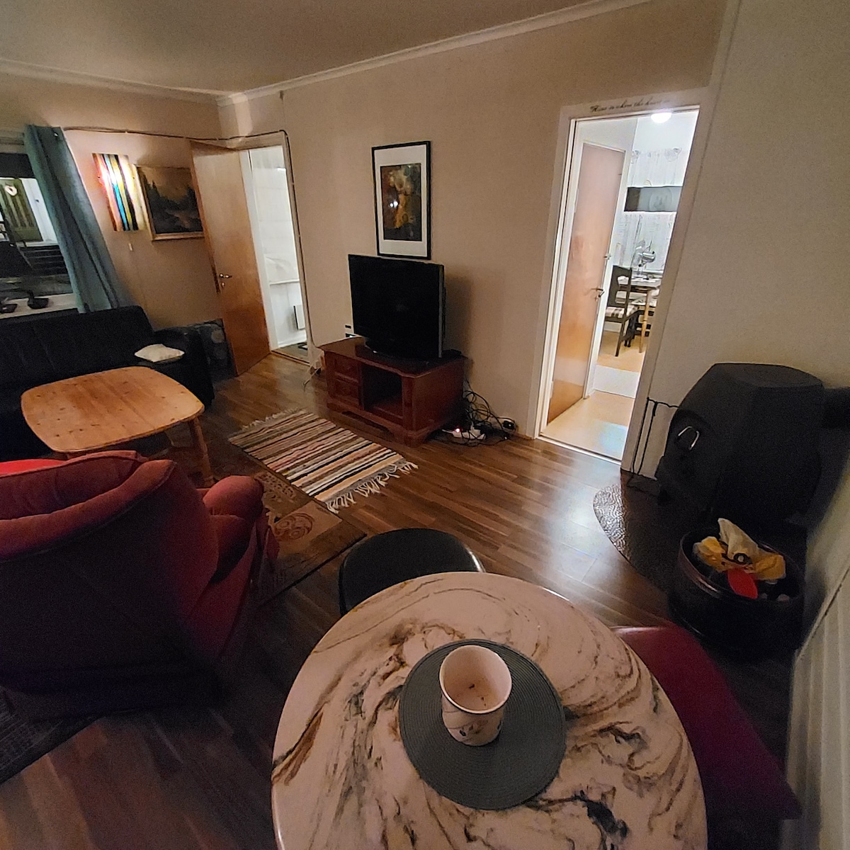 A living area features a television on a wooden stand, surrounded by a mix of furniture, including two distinct tables and a red armchair. Light wooden flooring is complemented by a colorful woven rug, while a doorway leads to a dining space in the background.