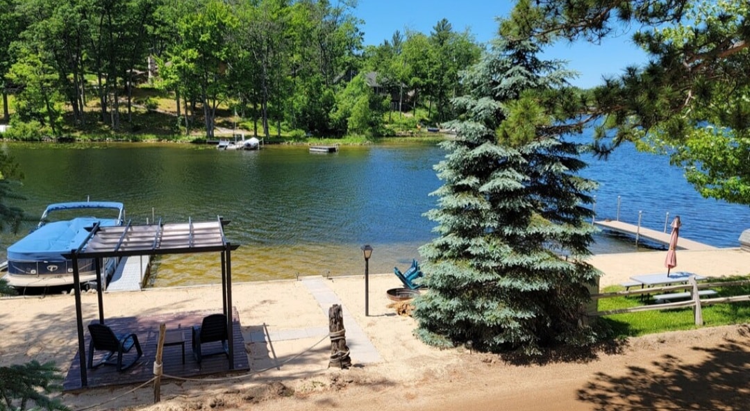 The tranquil lakefront is lined with lush greenery, showcasing a private dock and a patio area with seating. A shade structure provides rest from the sun, while a sandy area leads to the water's edge, inviting relaxation and outdoor activities.
