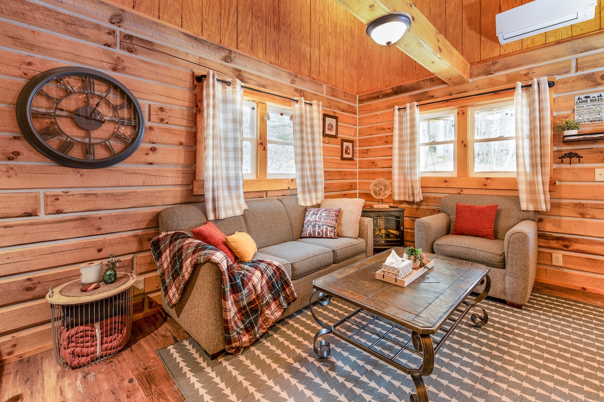 A warm living area features a cozy sofa adorned with a plaid blanket and decorative cushions. A rustic coffee table sits at the center, complemented by a vintage-style clock on the wall. Large windows allow natural light to illuminate the wood-paneled walls.