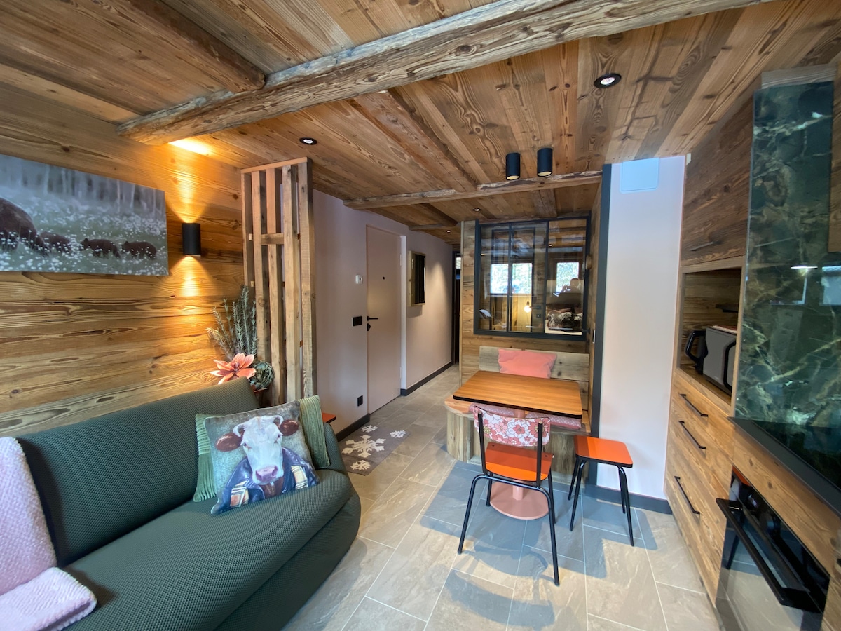 The living area features a green sofa adorned with a decorative pillow. A wooden dining table with four chairs is positioned adjacent to a kitchenette, highlighted by natural wood beams and stone accents throughout the space. Soft lighting creates a cozy atmosphere.