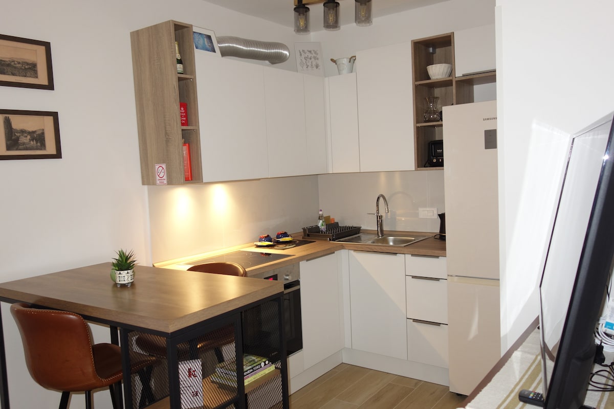 A modern kitchen features a blend of sleek white cabinets and wooden accents. The area is equipped with essential appliances, including a refrigerator and stove. A small dining table with two brown chairs is positioned beside a countertop, creating an inviting space for meal preparation and dining.