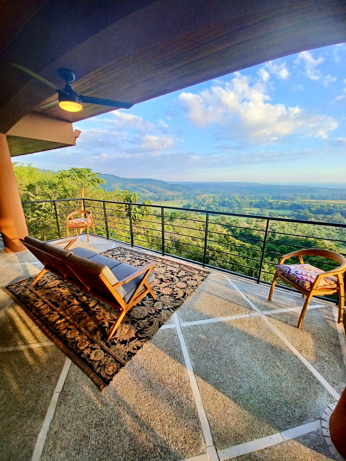 A spacious terrace is presented with a view over lush greenery, showcasing a mix of mountain and sky. Comfortable seating is arranged on a textured rug, complemented by a warm ceiling fan. The railing provides an unobstructed perspective of the surrounding landscape.