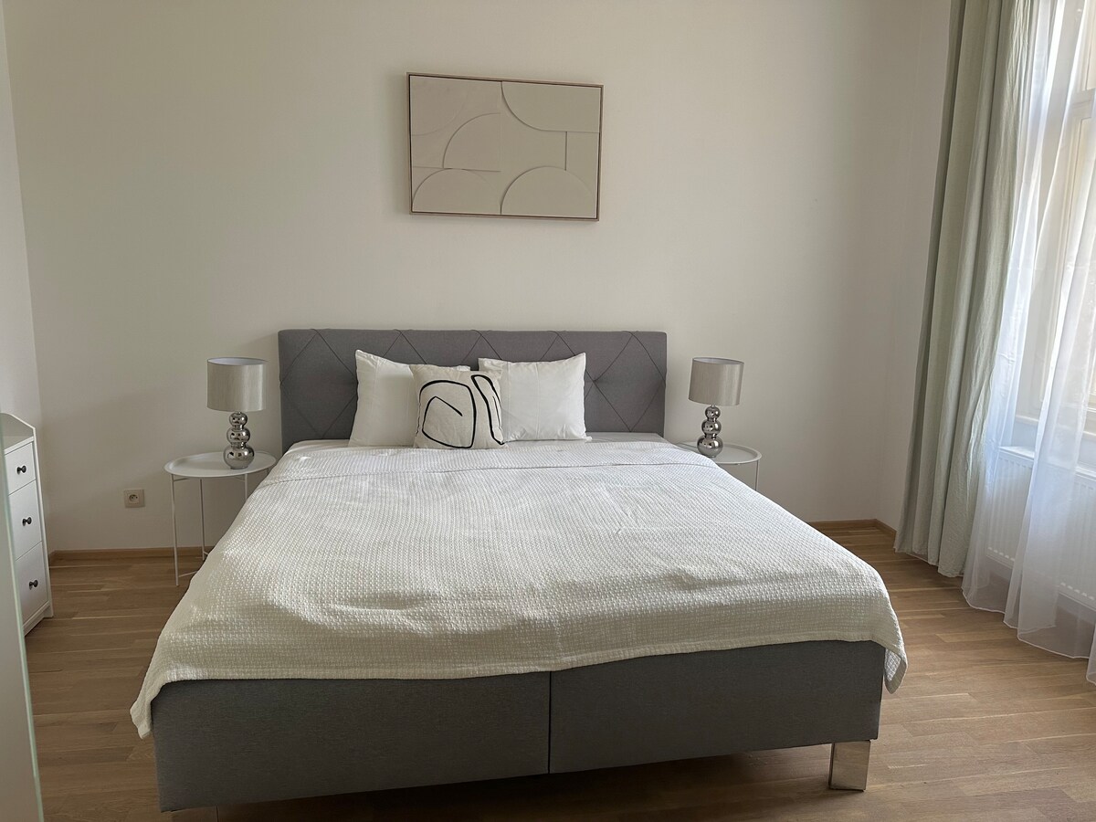 A spacious bedroom is presented with a large bed dressed in crisp white linens. Two bedside tables with lamps frame the bed, and natural light filters through sheer curtains, enhancing the room's bright and serene ambiance. A simple abstract wall art piece adds a modern touch.