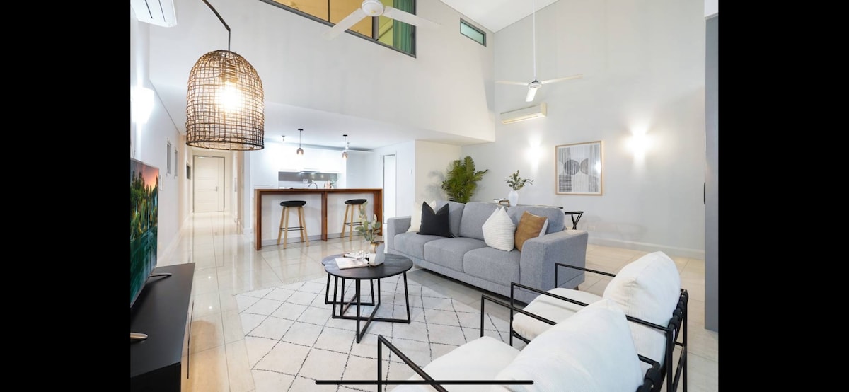 An open living area is illuminated by natural light, featuring a spacious grey sofa and contrasting black lounge chairs. A round coffee table sits atop a light-colored rug, while a kitchen with a bar counter and stools can be seen in the background.