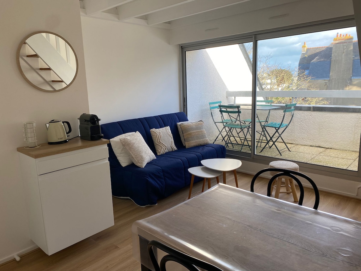 A modern living area features a blue convertible sofa, a dining table with black chairs, and an open kitchen setup. Large glass doors provide natural light and access to a terrace with outdoor seating. A round mirror is mounted on the white wall, adding a touch of style.