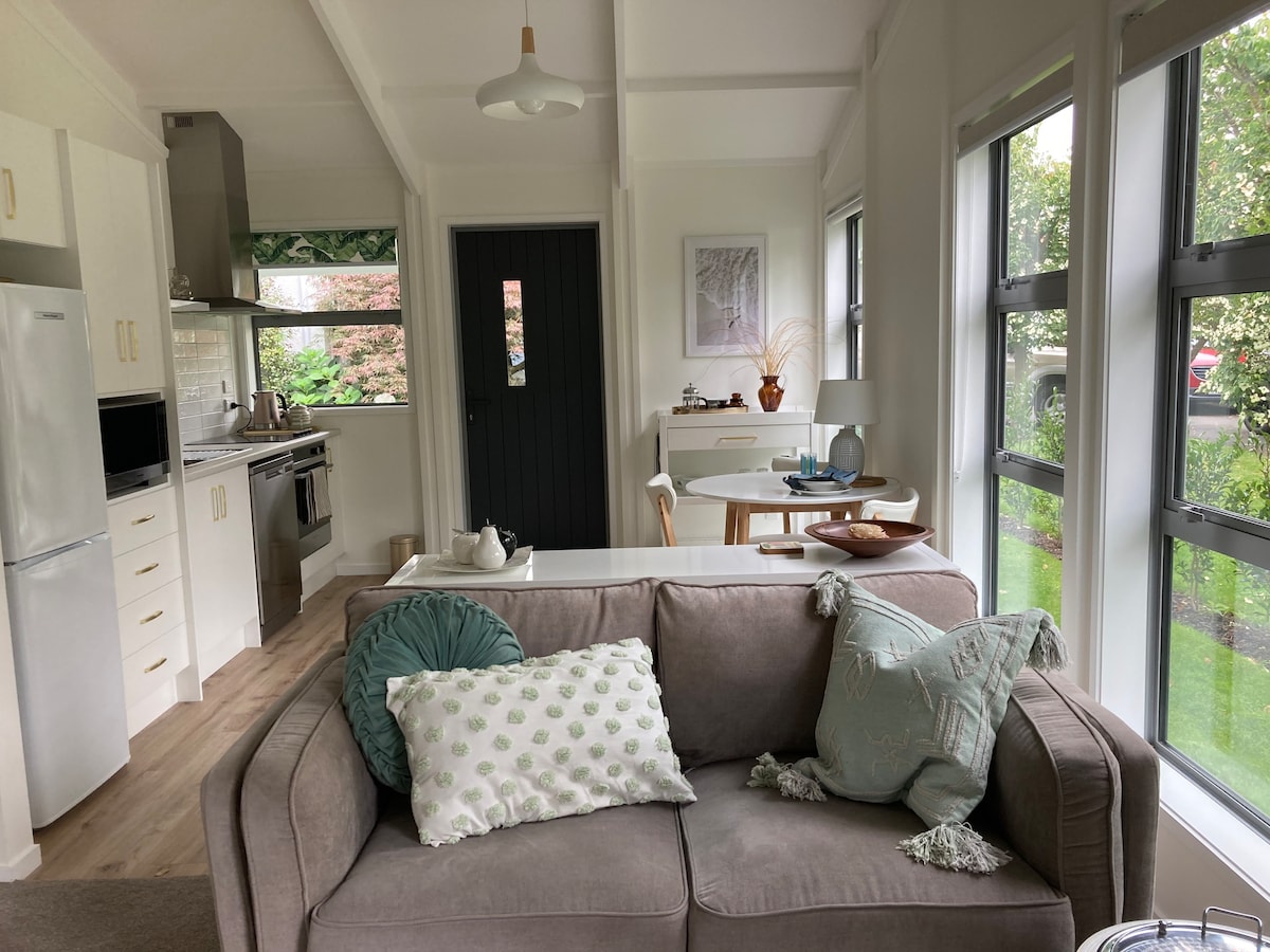 A cozy living area is furnished with a light-colored sofa adorned with patterned cushions. The space is illuminated by natural light streaming in through large windows, while a kitchenette is visible in the background. A dining table is positioned opposite the living area, creating a functional layout.
