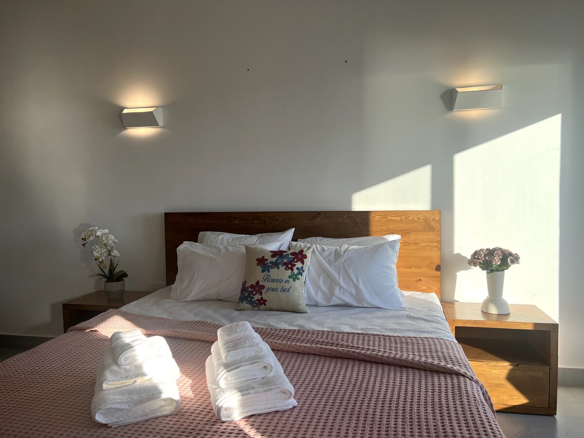 A cozy sleeping area features a wooden bed frame with white bedding and decorative pillows. Fluffy towels are neatly arranged at the foot of the bed. Soft wall lights illuminate the space, and a potted plant is placed on a bedside table, enhancing the serene atmosphere.