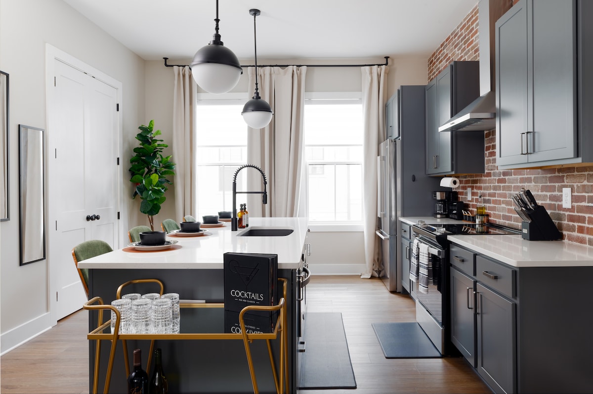 A modern kitchen is presented, featuring a spacious island with seating for four. The brick backsplash contrasts with grey cabinetry, while pendant lighting hangs above. Large windows allow natural light to illuminate the area, complemented by greenery and minimalistic décor for a functional yet stylish atmosphere.