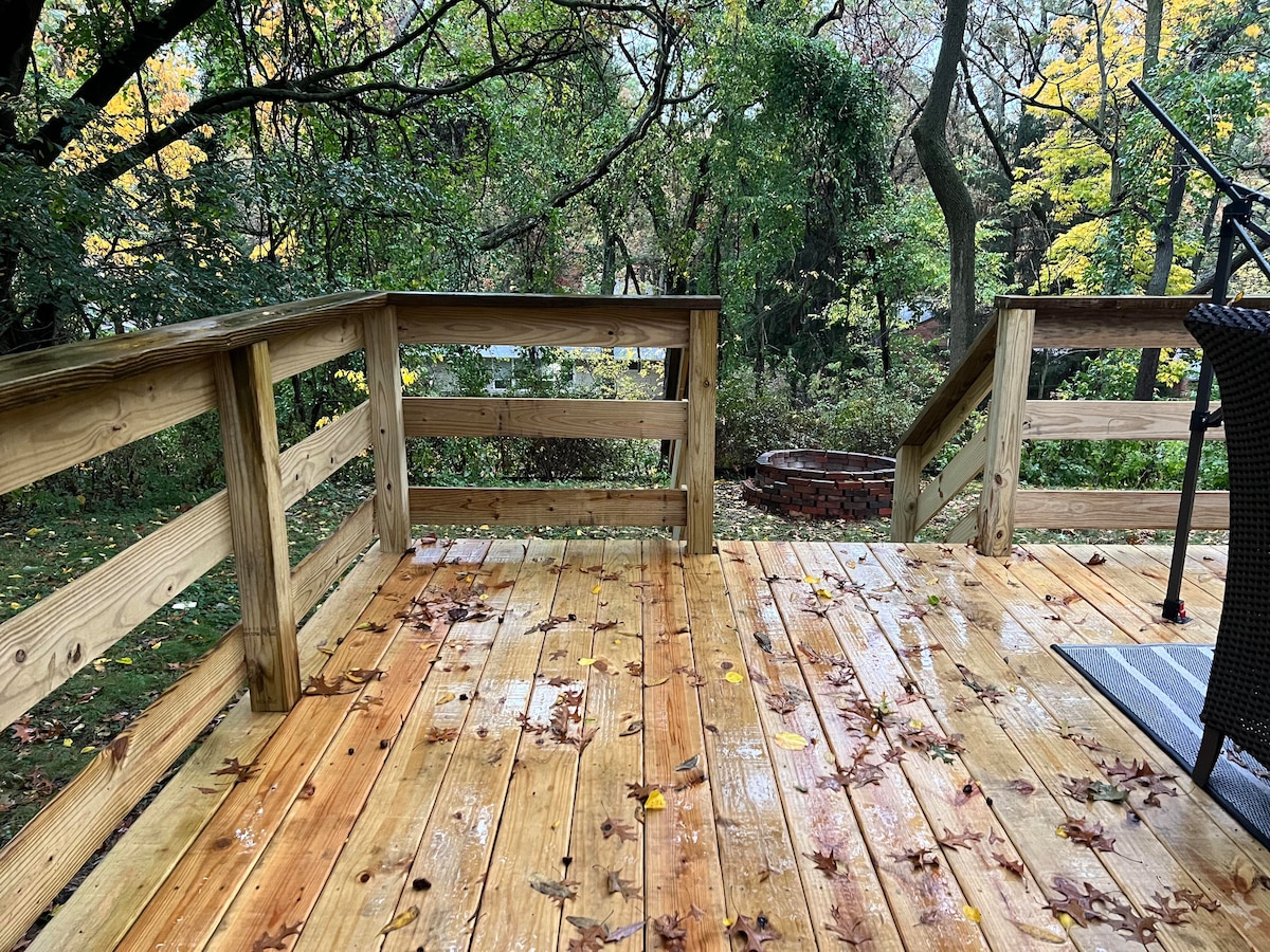 A wooden deck offers a spacious area for outdoor gatherings, surrounded by lush trees and foliage. A fire pit is visible in the background, and fallen leaves cover the surface, suggesting a serene, rustic atmosphere. The space invites relaxation and connection with nature.