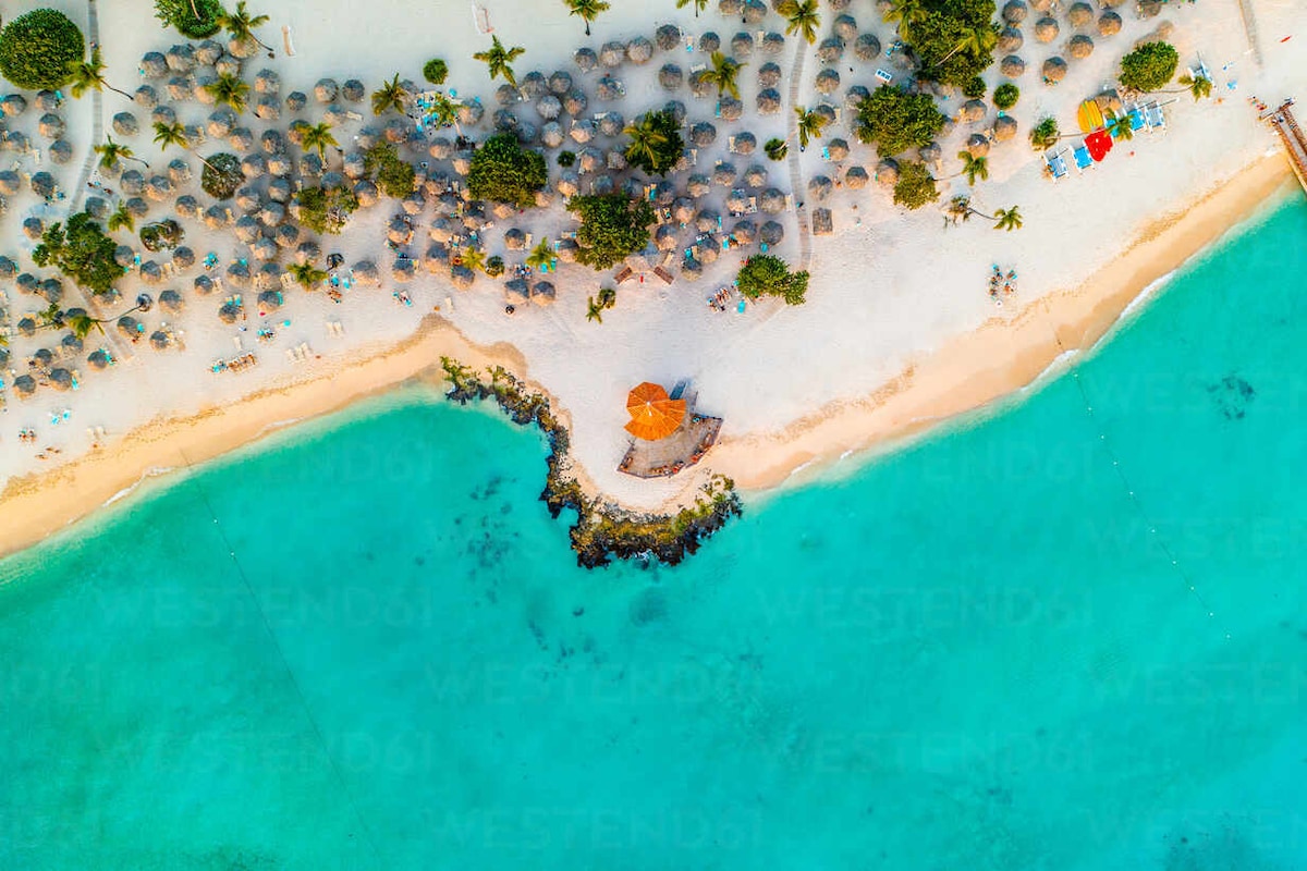 An aerial view captures a stretch of sandy beach bordered by clear turquoise waters. Umbrellas and lounge chairs dot the shoreline, while lush palm trees offer natural shade. A small structure is positioned at the water's edge, with rocky formations enhancing the coastal landscape.