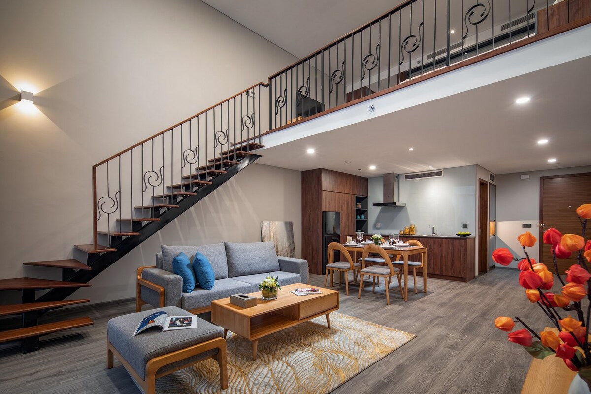 A spacious living area is captured, showcasing a comfortable sofa and a wooden coffee table adorned with decorative items. A dining table sits nearby, accompanied by modern kitchen appliances. A stylish staircase leads to the upper level, where light fills the space through large windows.