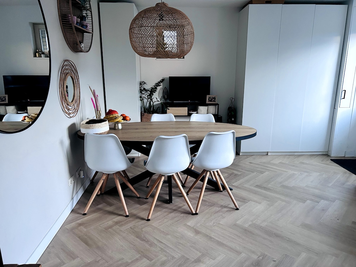 A dining area is featured, showcasing a large oval table surrounded by five modern, light-colored chairs with wooden legs. A woven pendant light hangs above the table, and a round mirror is mounted on the wall, reflecting the space's natural light.