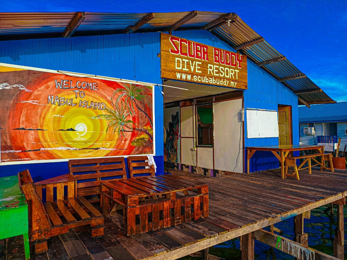 A wooden deck is visible in front of a colorful mural that welcomes guests to Mabul Island, featuring sunset imagery and palm trees. A sign with the name 'Scuba Buddy Dive Resort' is prominently displayed. Rustic wooden furniture is arranged for outdoor seating.