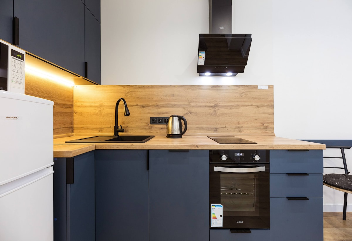 A modern kitchen featuring sleek dark cabinetry and a wood-accented backsplash. Stainless steel appliances, including an oven and a kettle, are positioned on a light countertop. Soft under-cabinet lighting enhances the workspace, while a white refrigerator stands discreetly nearby.