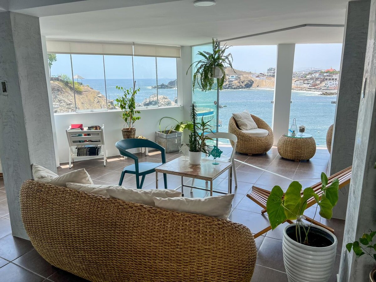 A spacious living area is filled with natural light, featuring comfortable woven furniture and a glass table. Large windows offer unobstructed views of the ocean, and local greenery enhances the airy atmosphere. A hammock invites relaxation in this welcoming coastal retreat.