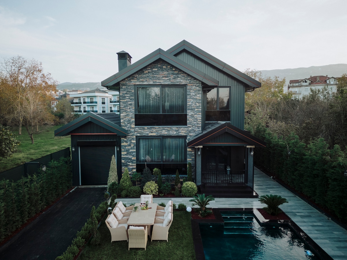 A modern two-story villa features large windows and stone accents. A covered patio area includes a dining table surrounded by wicker chairs. A pathway leads to a small pool set amidst landscaped greenery, enhancing the outdoor living space.