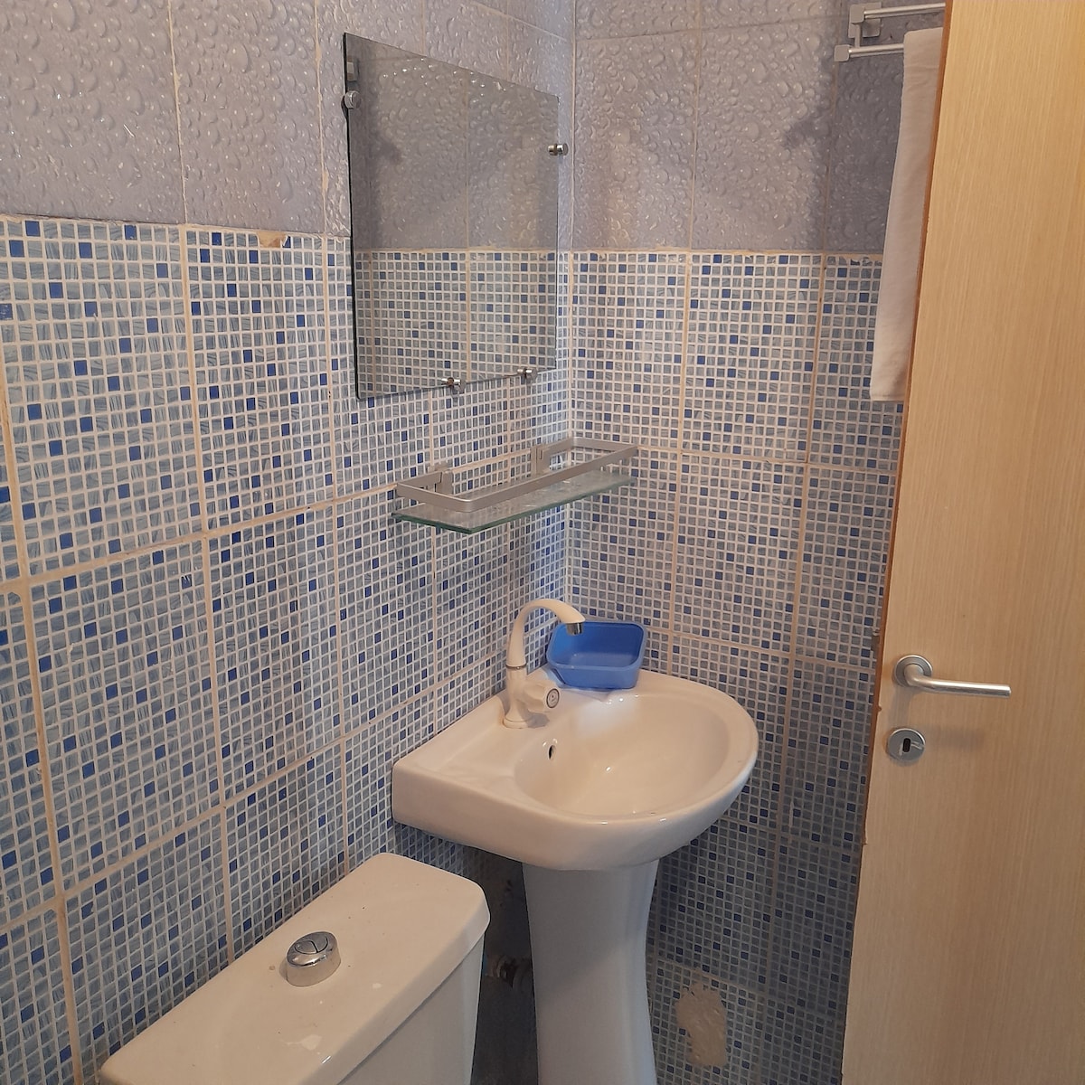 The bathroom features mosaic-tiled walls in shades of blue, creating a serene environment. A modern sink is positioned beside a compact toilet, with a mirror mounted above the sink. A small shelf is attached to the wall for toiletries.