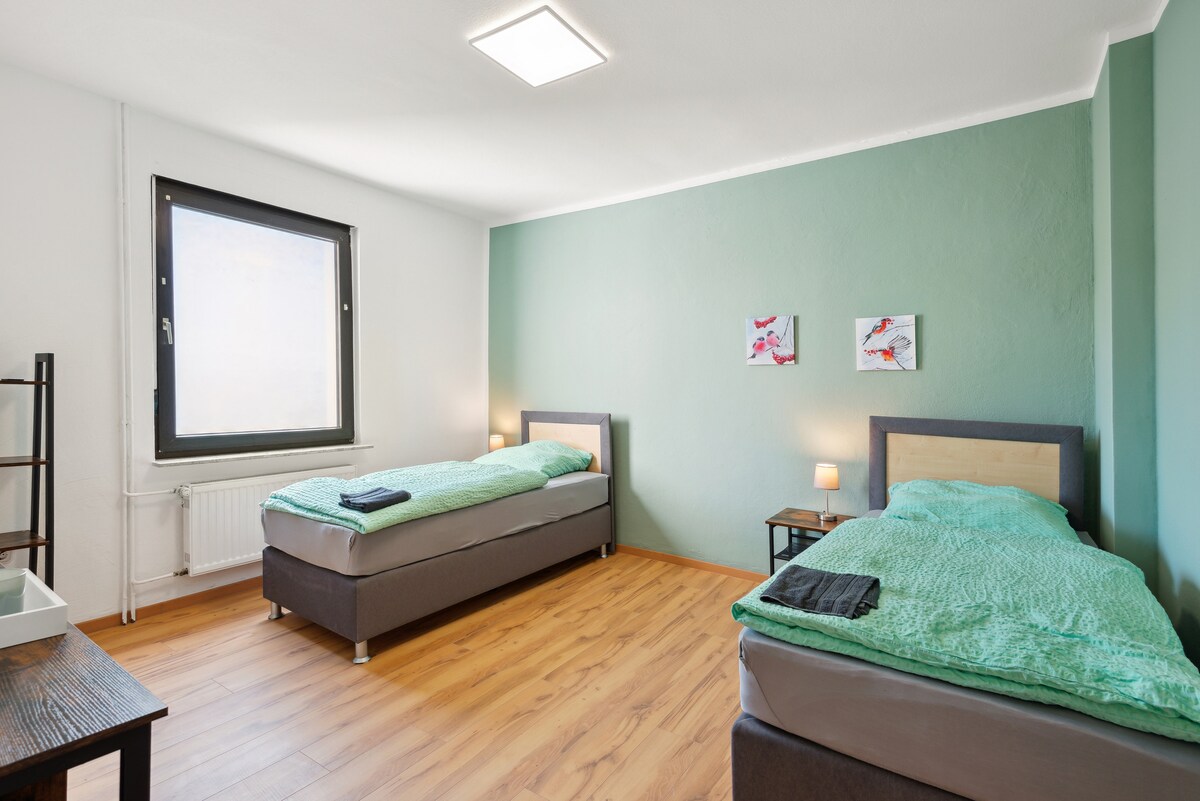 Ფართო საძინებელი ორი ერთადგილიანი საწოლით, მწვანე აქცენტის კედლით და მარტივი დეკორაციით. Იდეალურია დასასვენებელი ატმოსფეროსთვის.