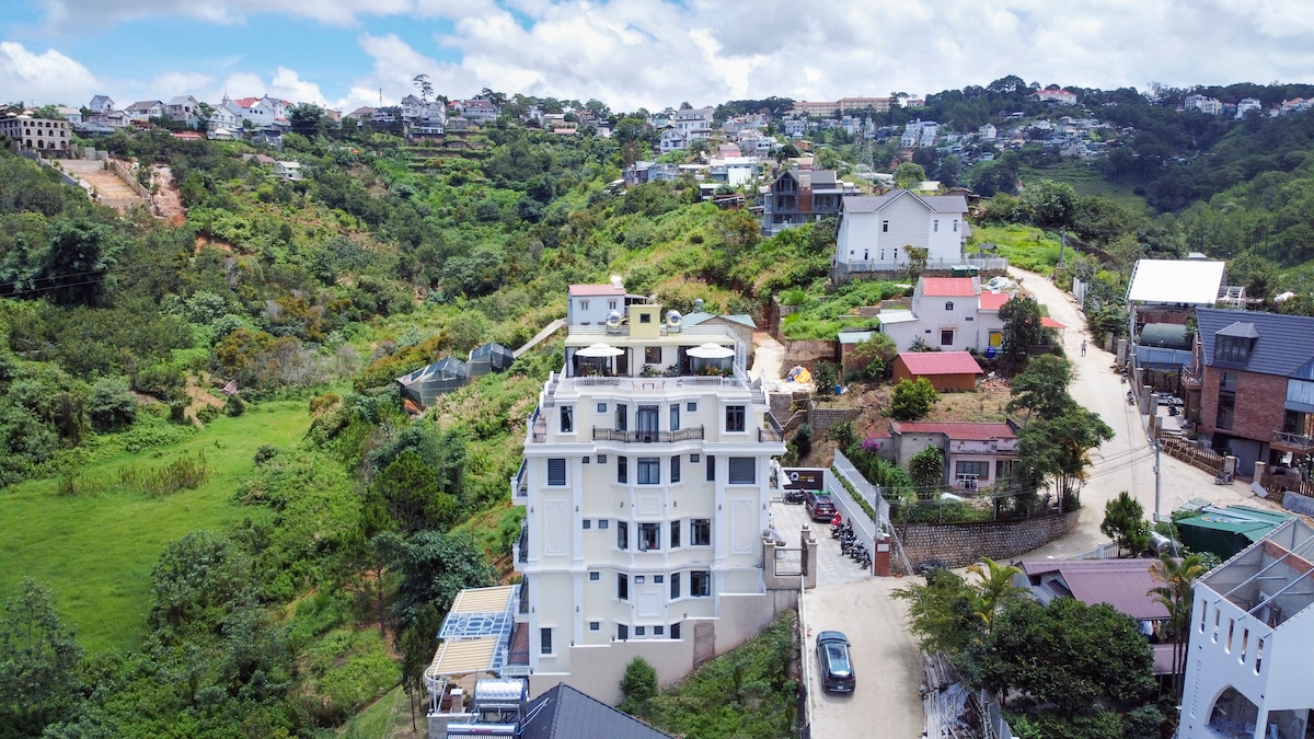 A modern multi-story building is situated amidst lush green hills, showcasing a scenic view of the surrounding landscape. Nearby structures and a winding road are visible in the foreground. The setting emphasizes the property's location in a tranquil valley.