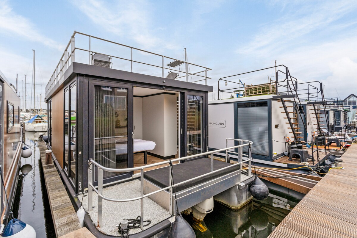 The waterloft features a modern and sleek design with large glass doors leading to a spacious deck area. The deck is surrounded by calm waters, offering direct access to the exterior while providing views of neighboring houseboats and the marina.