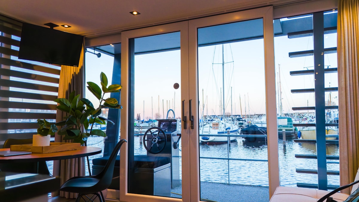 Large glass doors provide a view of the marina, showcasing numerous boats docked in the water. Natural light fills the space, illuminating a dining area with a wooden table and black chairs. Indoor plants are placed on the table, adding a touch of greenery.